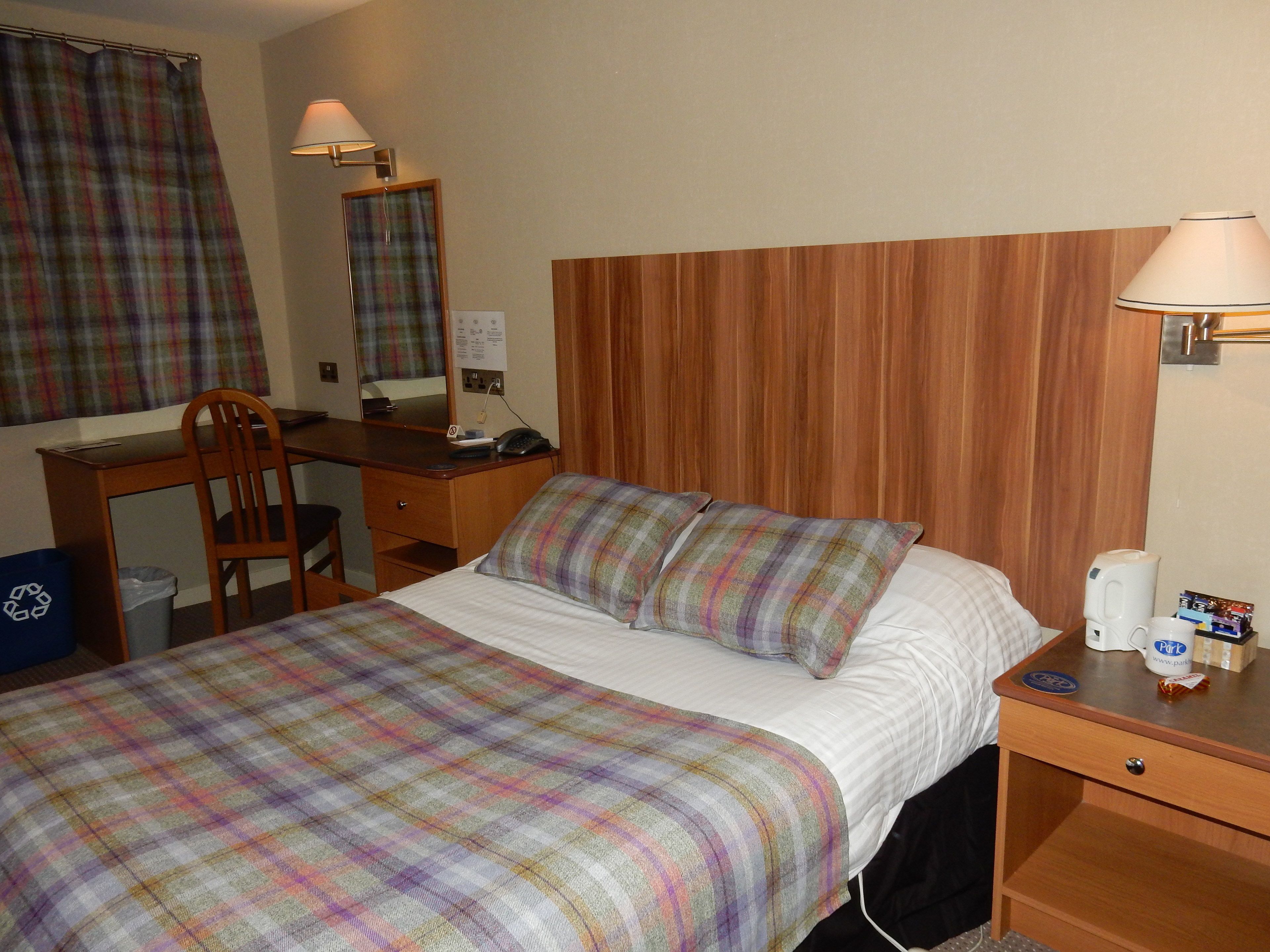 Habitación de hotel con cama, escritorio, silla, espejo, lámpara y mesita de noche con teléfono y taza.