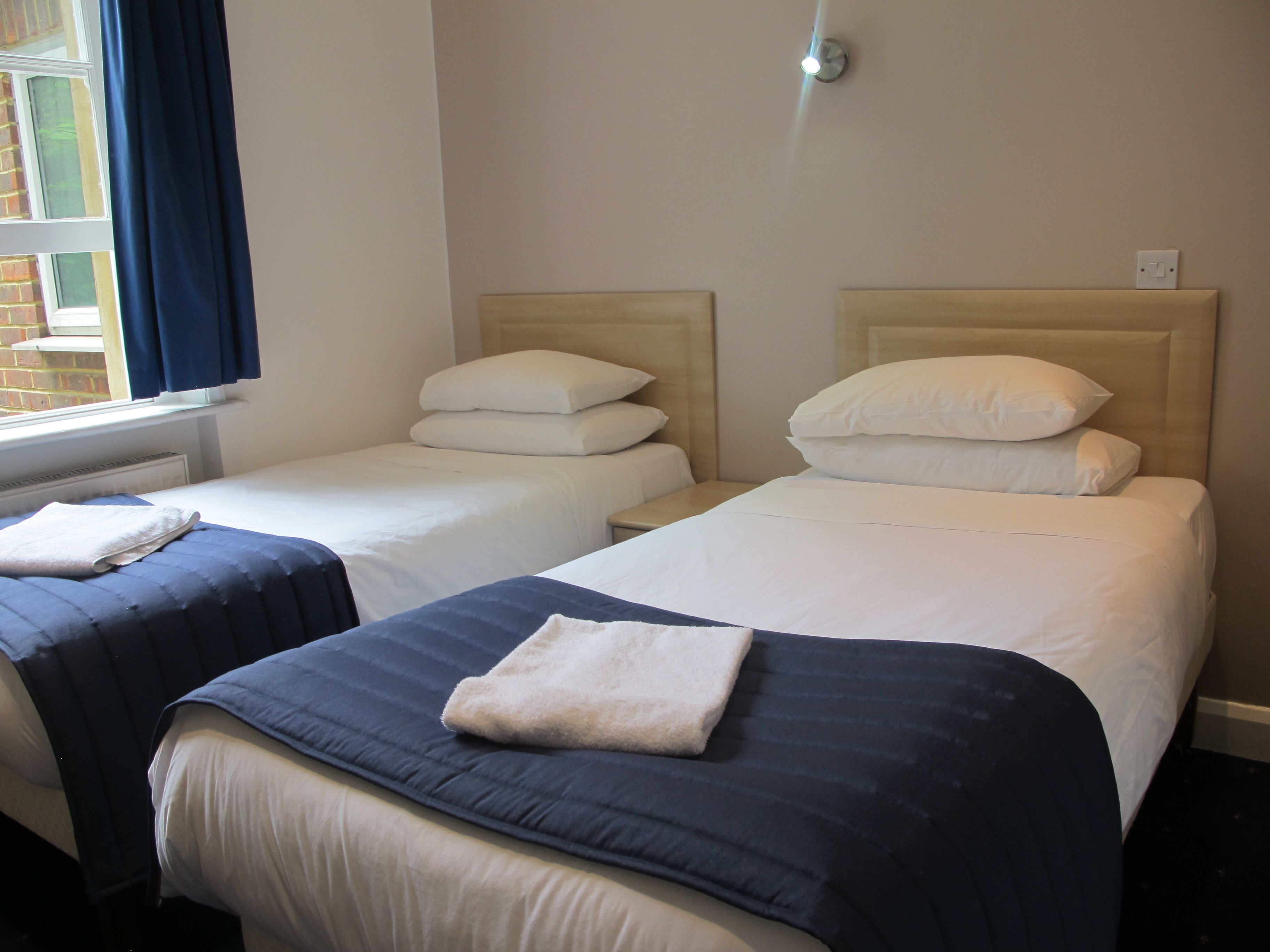 A hotel room with two single beds, each with a headboard, a window with curtains, and a wall-mounted light fixture.