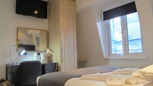 Hotellihuone, jossa on sänky, työpöytä lampun kanssa, peili, hiustenkuivaaja ja ikkuna, josta on näkymä rakennuksiin.
