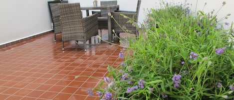 An outdoor patio with a tiled floor, wicker furniture, and a planter with purple flowers.