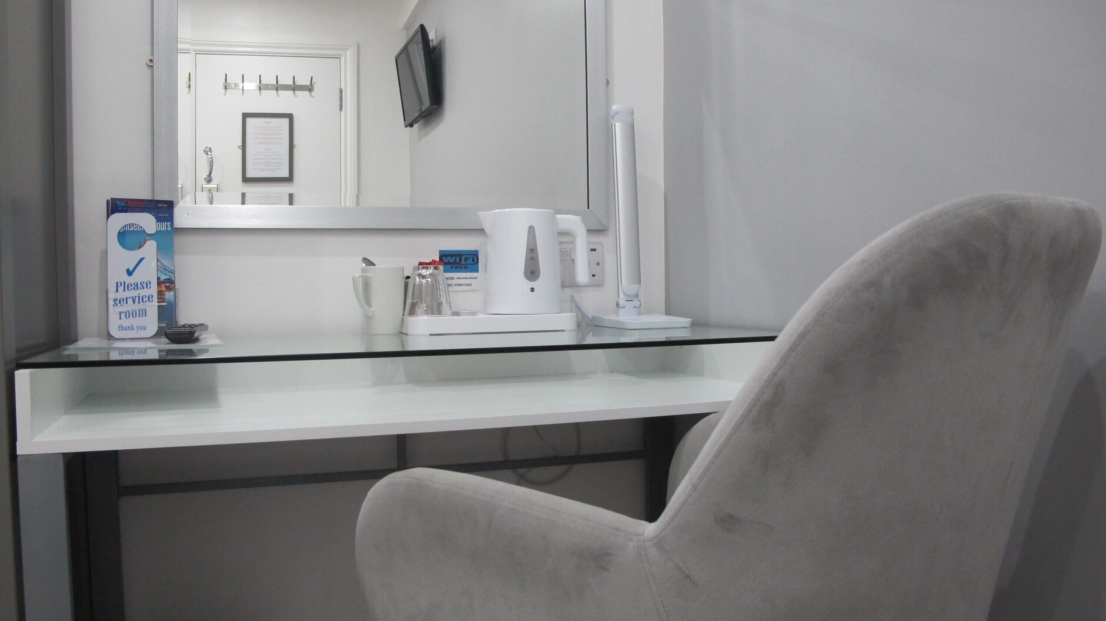 A hotel room with a white desk, a white chair, a coffee maker, a WiFi sign, and a TV mounted on the wall.