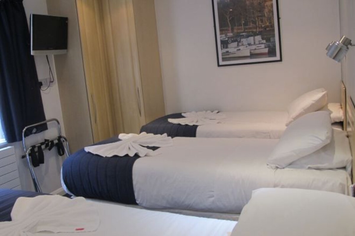 Ein Hotelzimmer mit zwei Betten, einem an der Wand befestigten Fernseher, einem Fenster mit Vorhängen und einem gerahmten Bild.