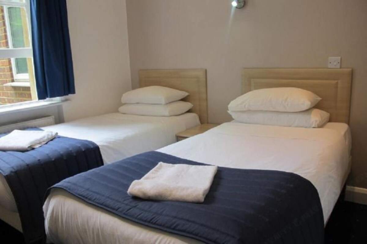 Ein Hotelzimmer mit zwei Einzelbetten, jeweils mit Kopfteil, einem Fenster mit Vorhängen und einer Wandleuchte.