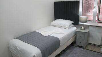 A neatly made bed with white and gray bedding, a bedside table with a lamp, and a window with curtains.