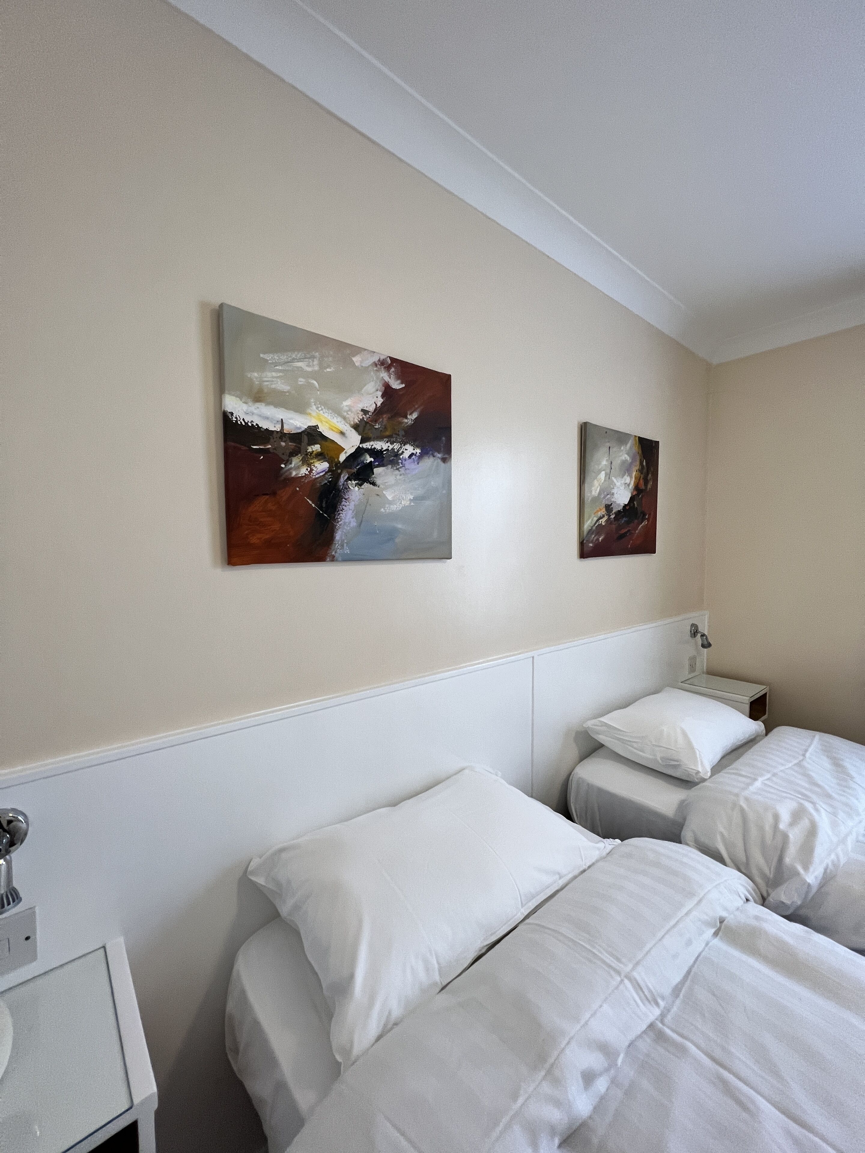 A hotel room with two single beds, a nightstand with a lamp, and two abstract paintings on the wall.