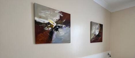 A hotel room with two single beds, a nightstand with a lamp, and two abstract paintings on the wall.