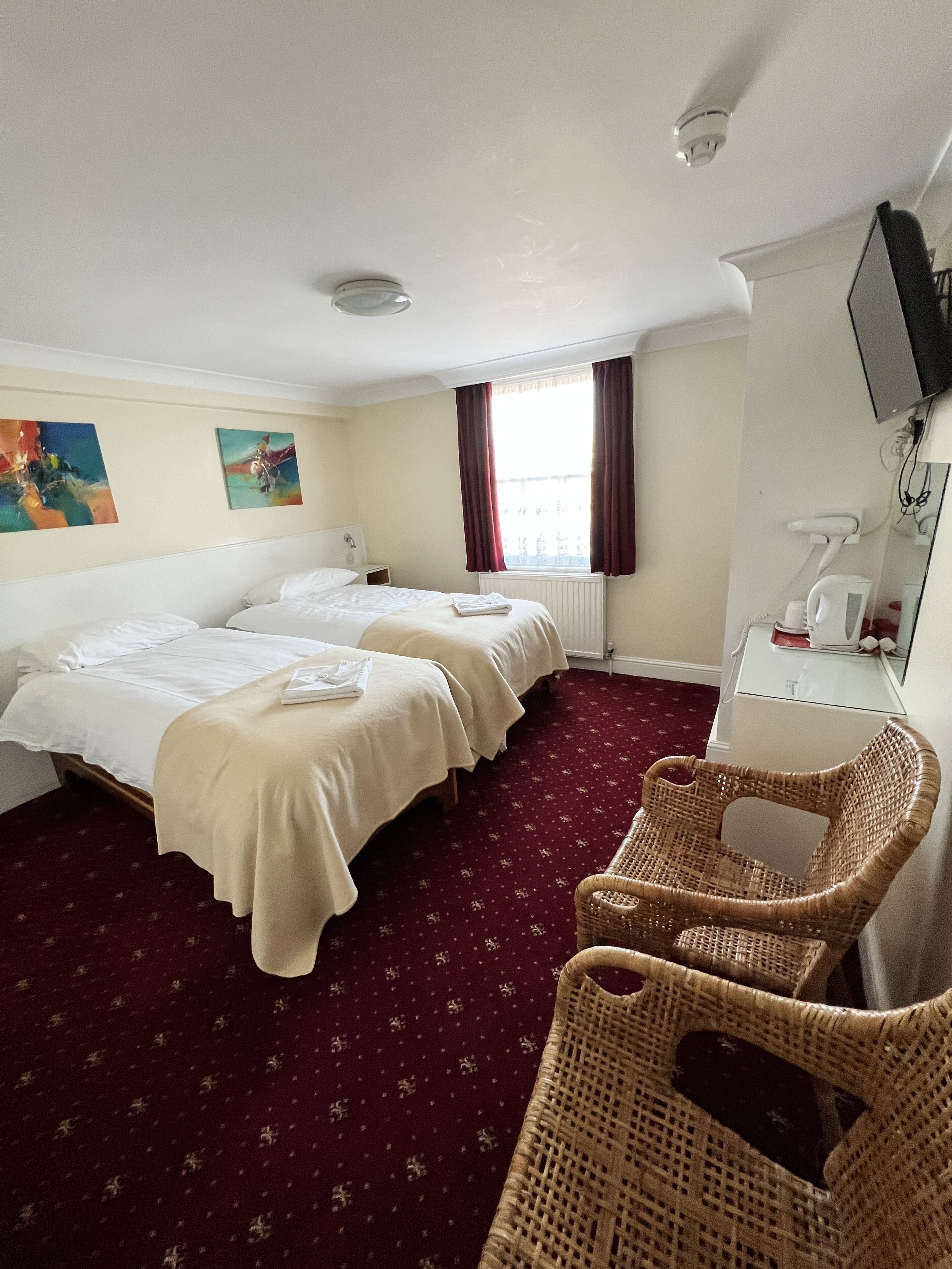 A hotel room with two beds, a red carpet, a wicker chair, a desk, and a television on the wall.