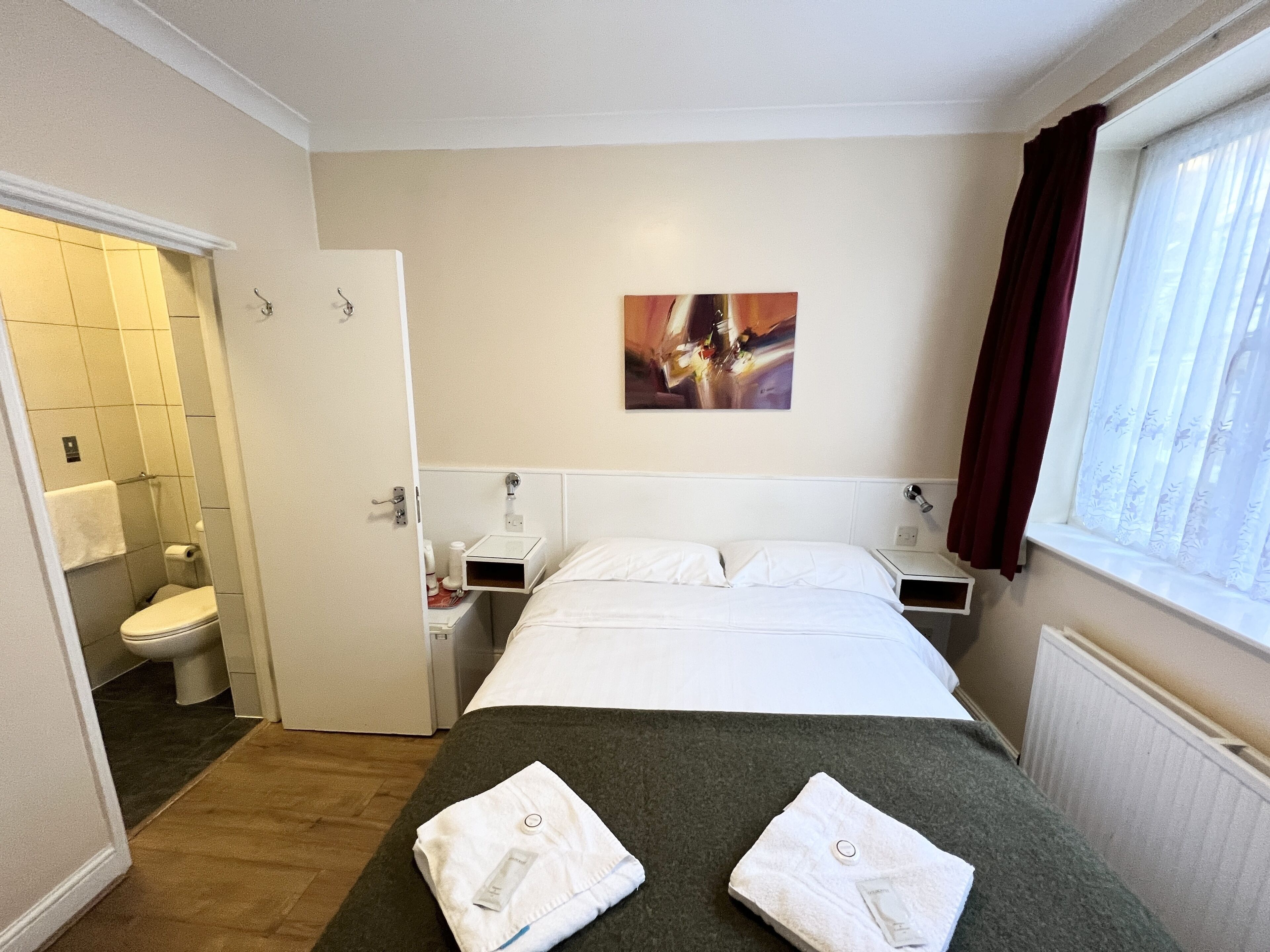 A hotel room with a bed, a nightstand, a bedside table, a window with curtains, a painting on the wall, and a bathroom with a toilet and towel rack.