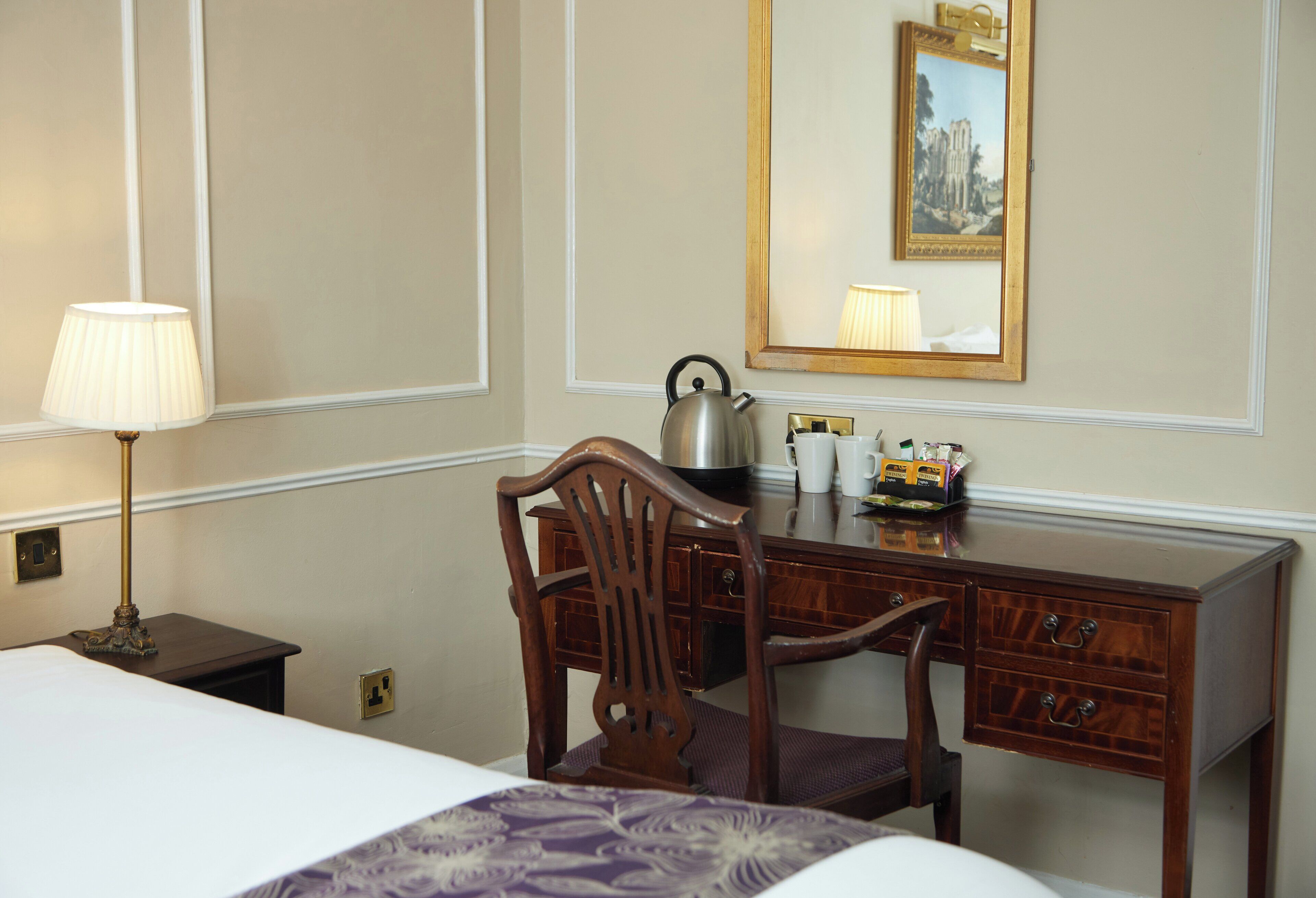 Habitación de hotel con cama, escritorio con hervidor, silla, lámpara y espejo con un cuadro.