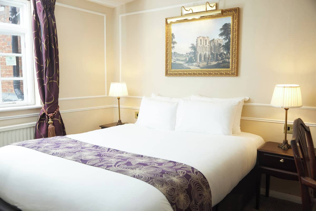 A neatly made bed with a purple and white bedspread, two bedside lamps, a framed painting on the wall, and a window with curtains.