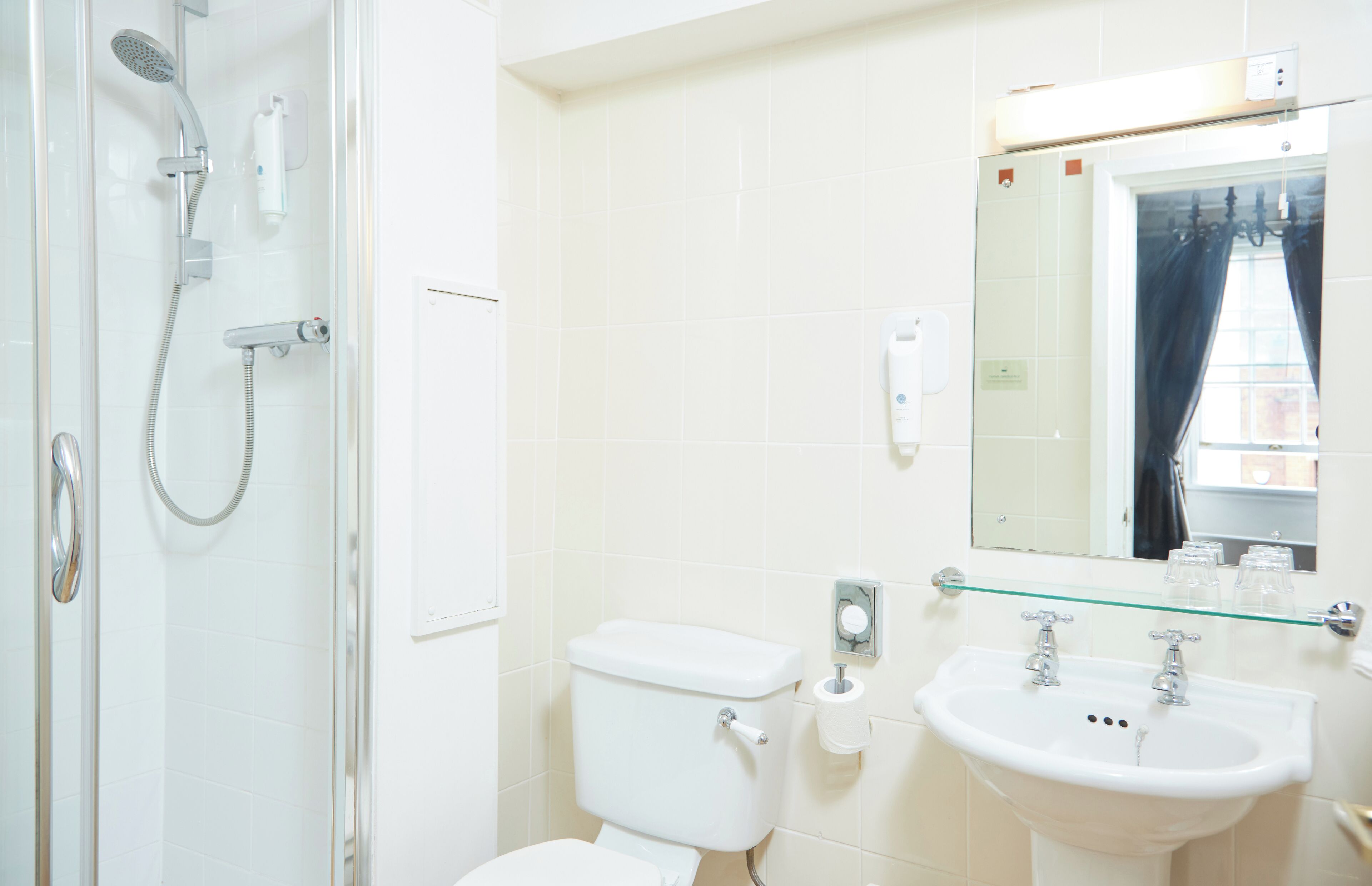 A bathroom with a glass shower enclosure, a toilet, a sink, and a mirror with a glass shelf above it.