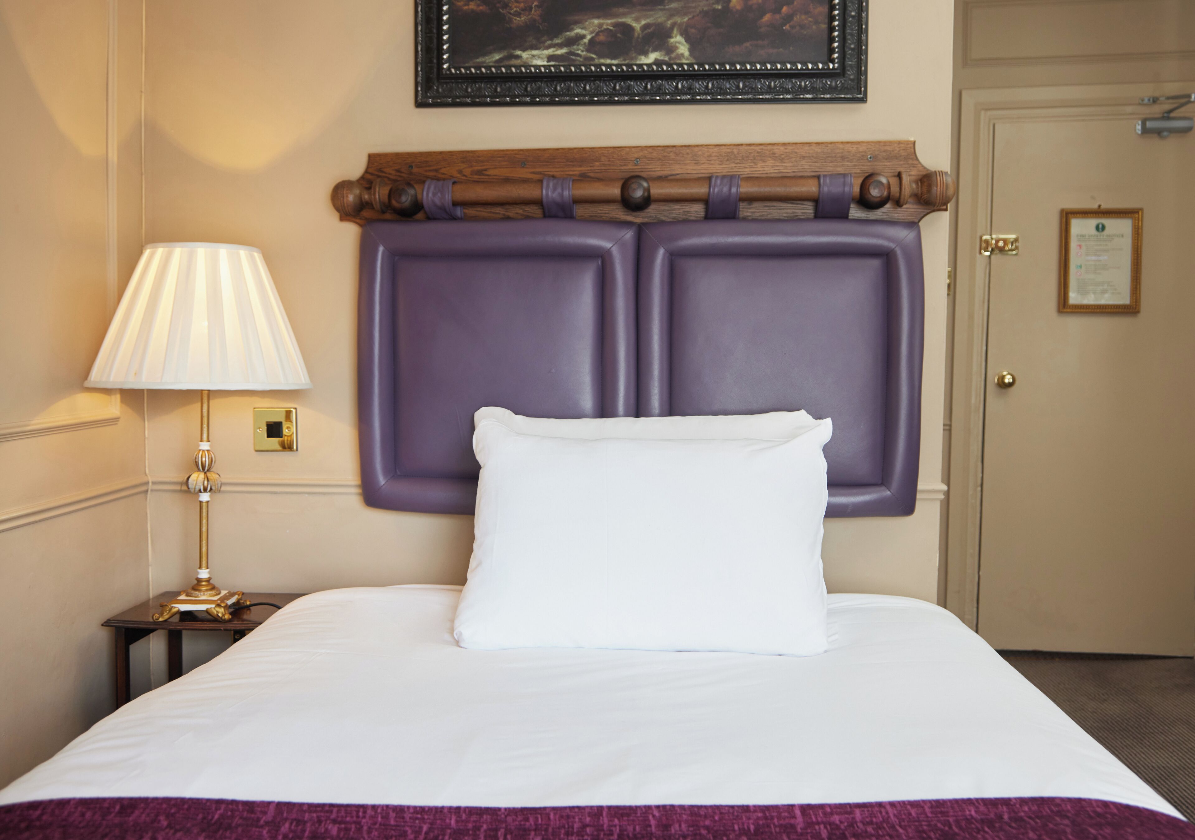 A hotel room with a bed, a nightstand with a lamp, and a door with a sign.