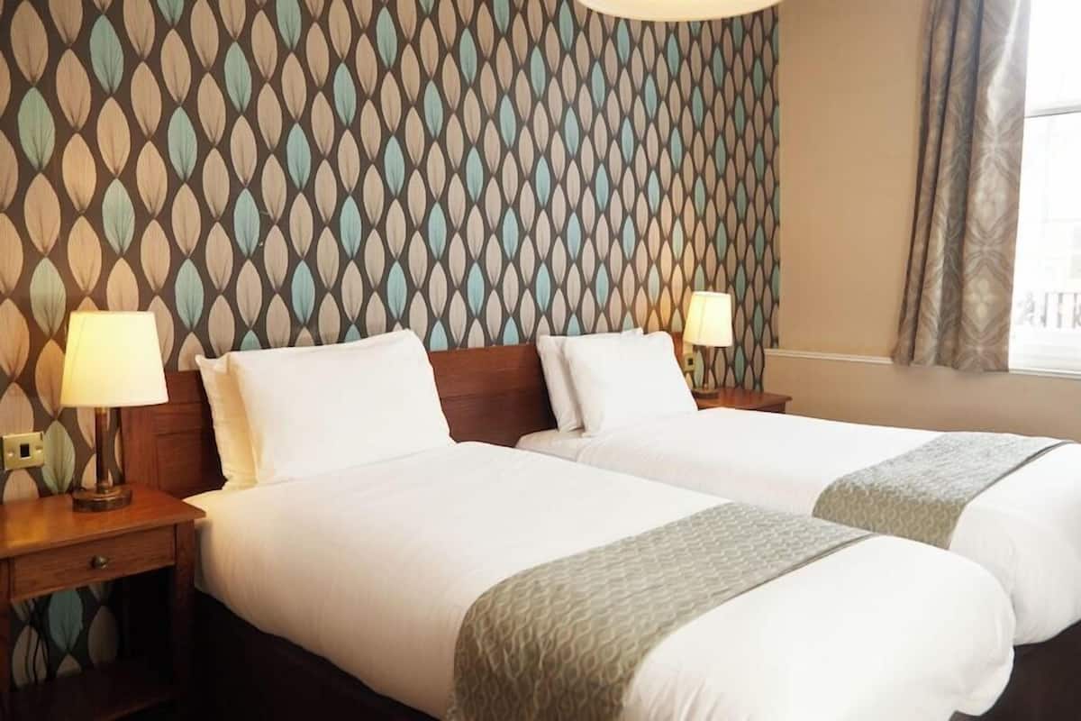 A hotel room with two beds, wooden headboards, bedside lamps, a patterned wallpaper, and a window with curtains.