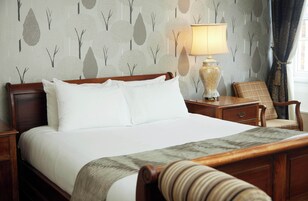 Una cama bien hecha con sábanas blancas, cabecera de madera y una mesita de noche con lámpara.