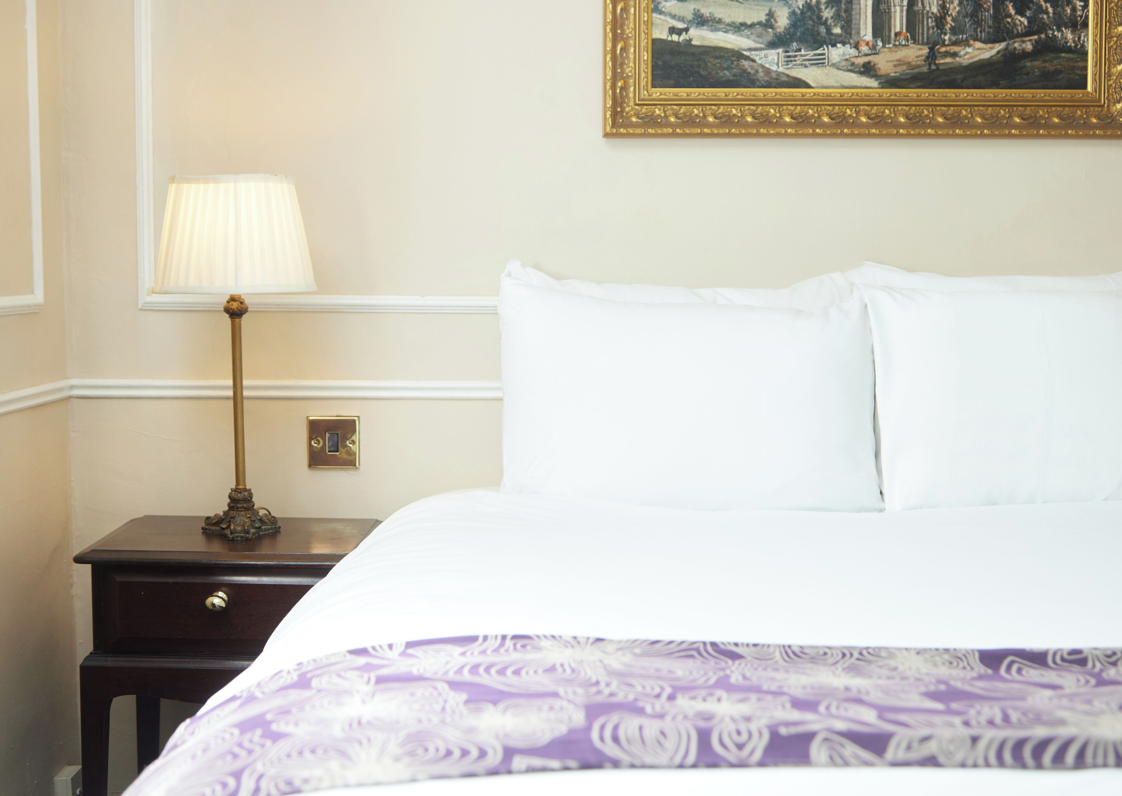 A hotel room with a bed, a nightstand with a lamp, a painting on the wall, and a door with a handle.