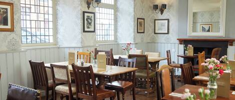 A dining area with wooden tables and chairs, framed pictures on the walls, and a large mirror above the fireplace.