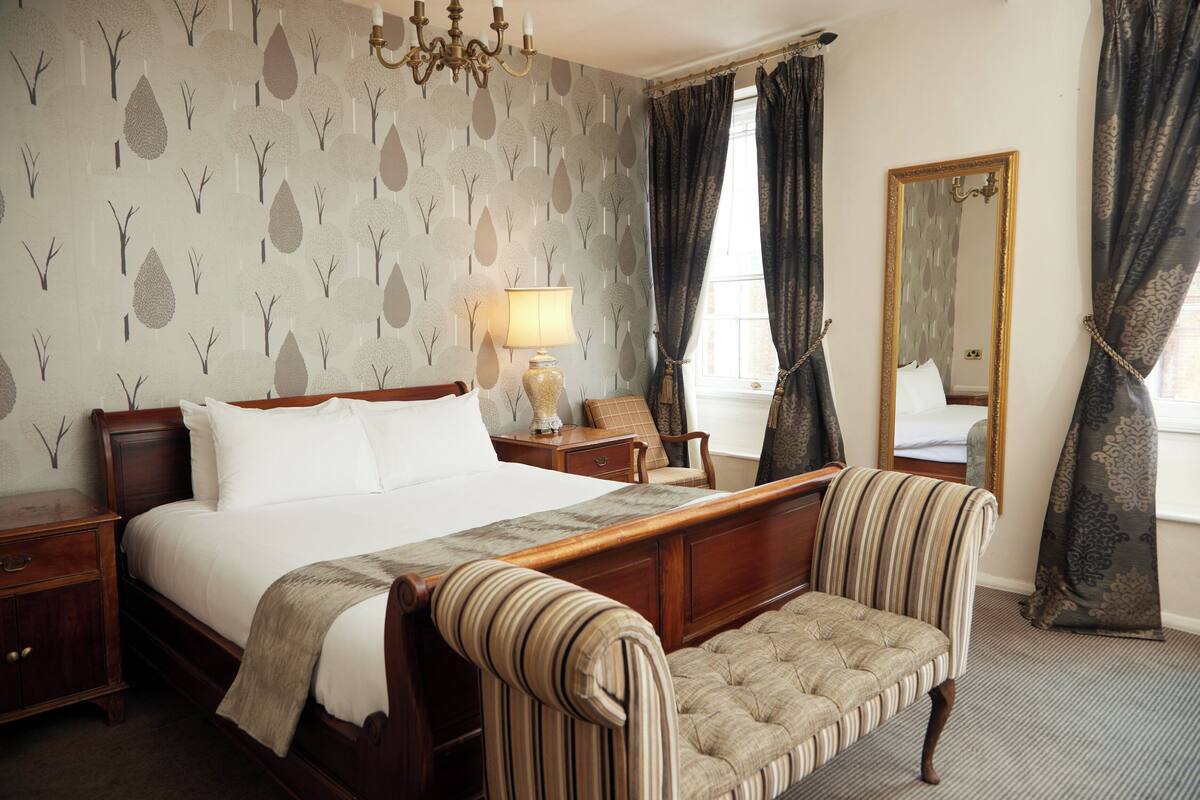 A neatly arranged bedroom with a large bed, a bedside table, a lamp, a chair, a mirror, and a window with curtains.