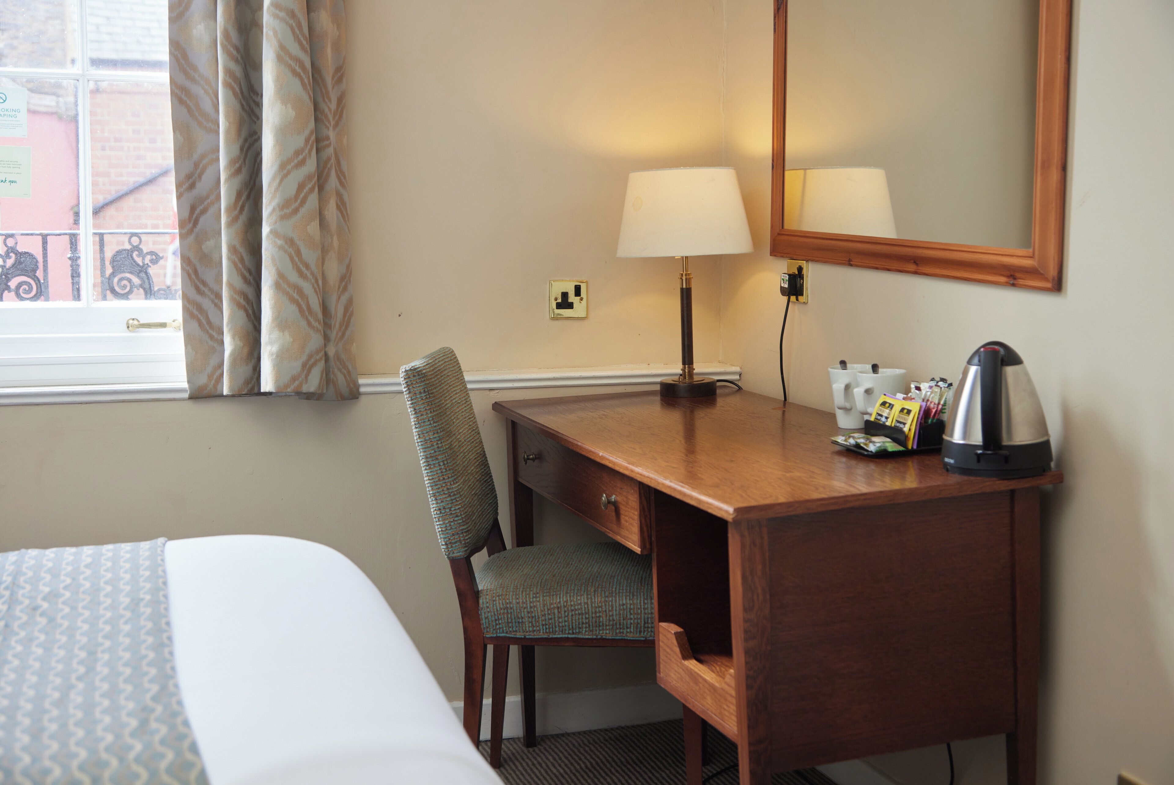 Une chambre d’hôtel comprenant un lit, un bureau en bois, une chaise, une lampe, une bouilloire et un miroir.