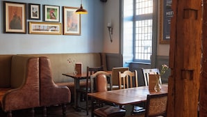 A cozy restaurant interior with a wooden table, cushioned seating, framed artwork on the wall, and a window with a view. - Cock Hotel Stony Stratford by Greene King Inns (Milton Keynes)