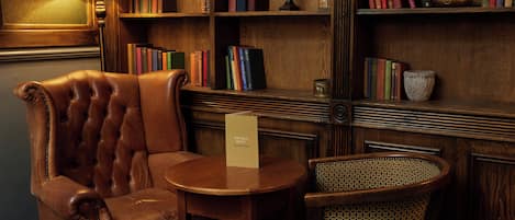 A leather armchair, a small round table, and a wicker chair in a library setting with bookshelves and a lamp.