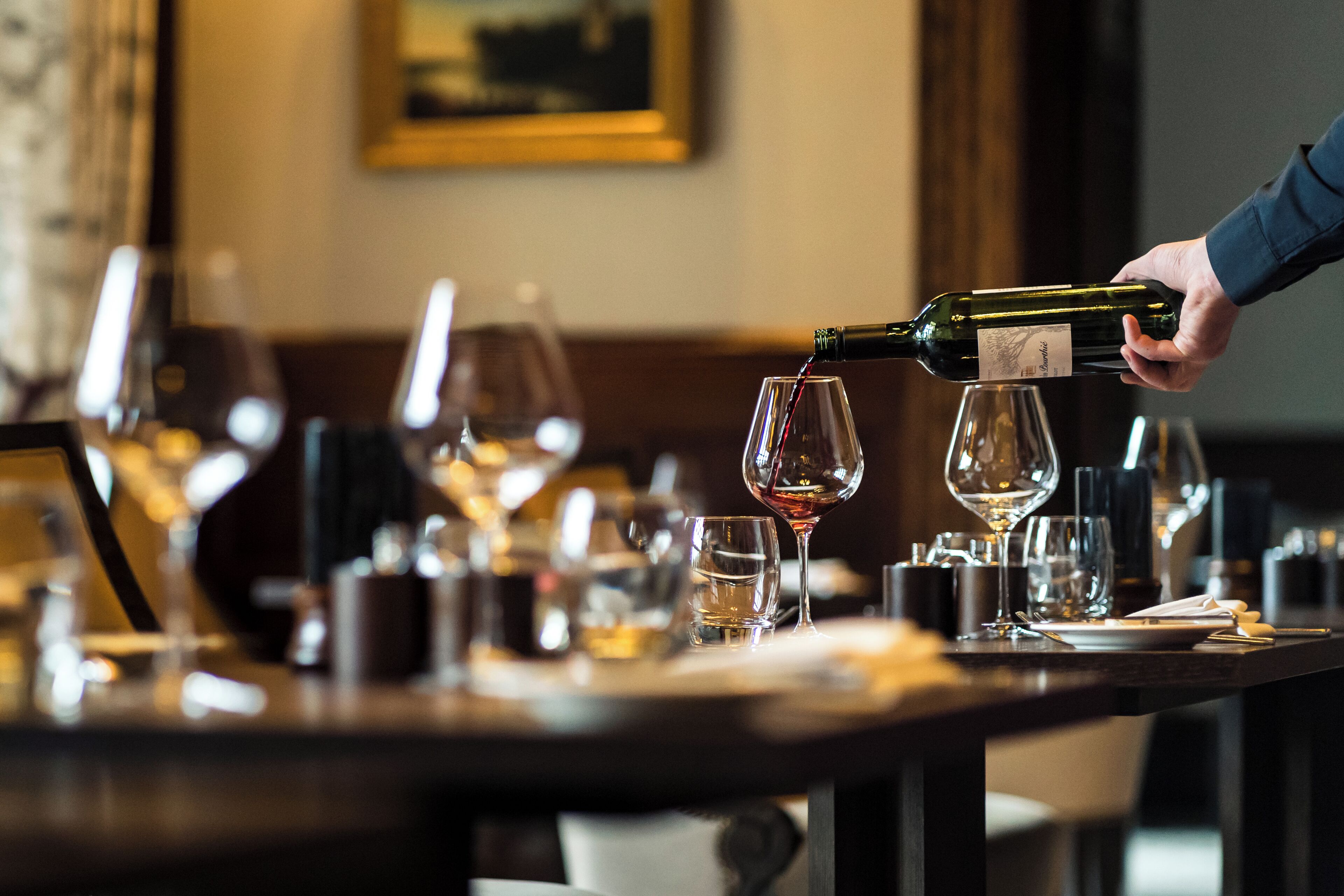 En person hælder vin i et glas i en bar med flere glas og flasker.