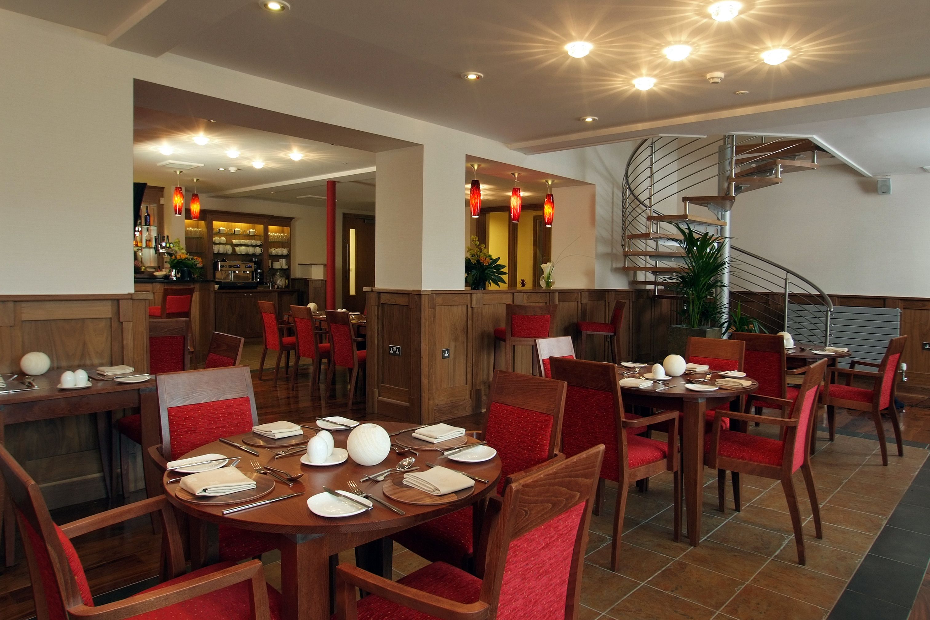 A dining area with round tables set for four, red upholstered chairs, and a spiral staircase in the background.