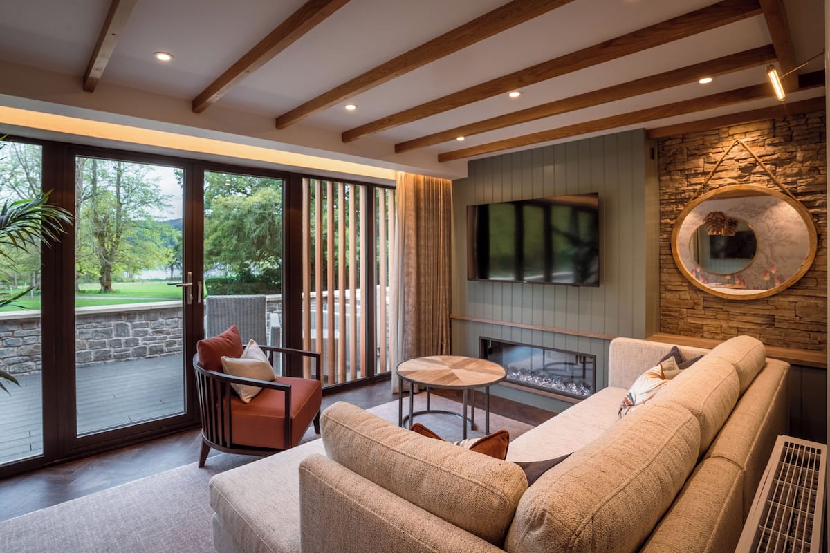 A modern living room with a sofa, a chair, a small table, and a fireplace. There is a large window with a view of greenery outside.