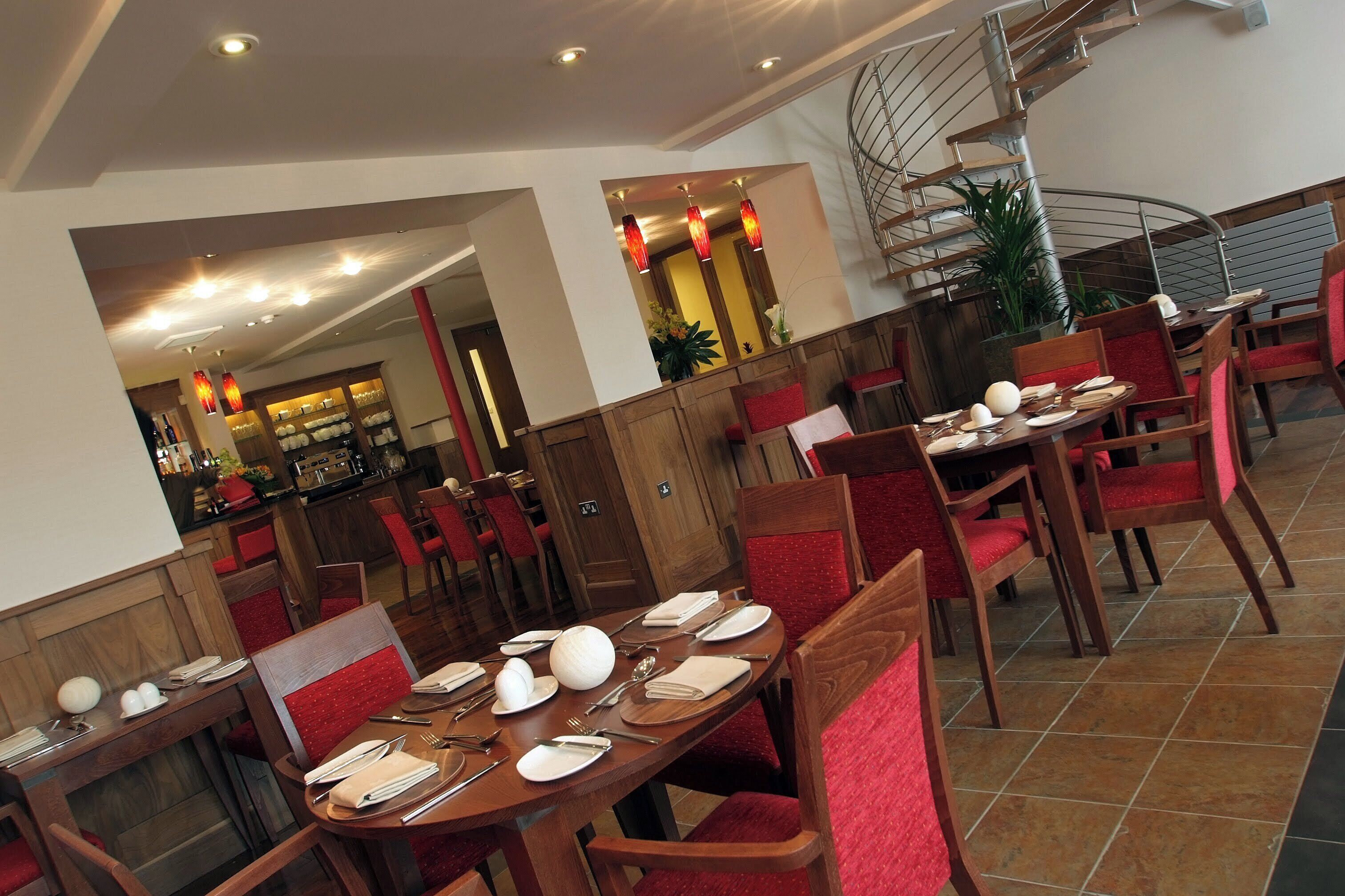 A dining area with wooden tables and red upholstered chairs, a spiral staircase, and a bar area in the background.