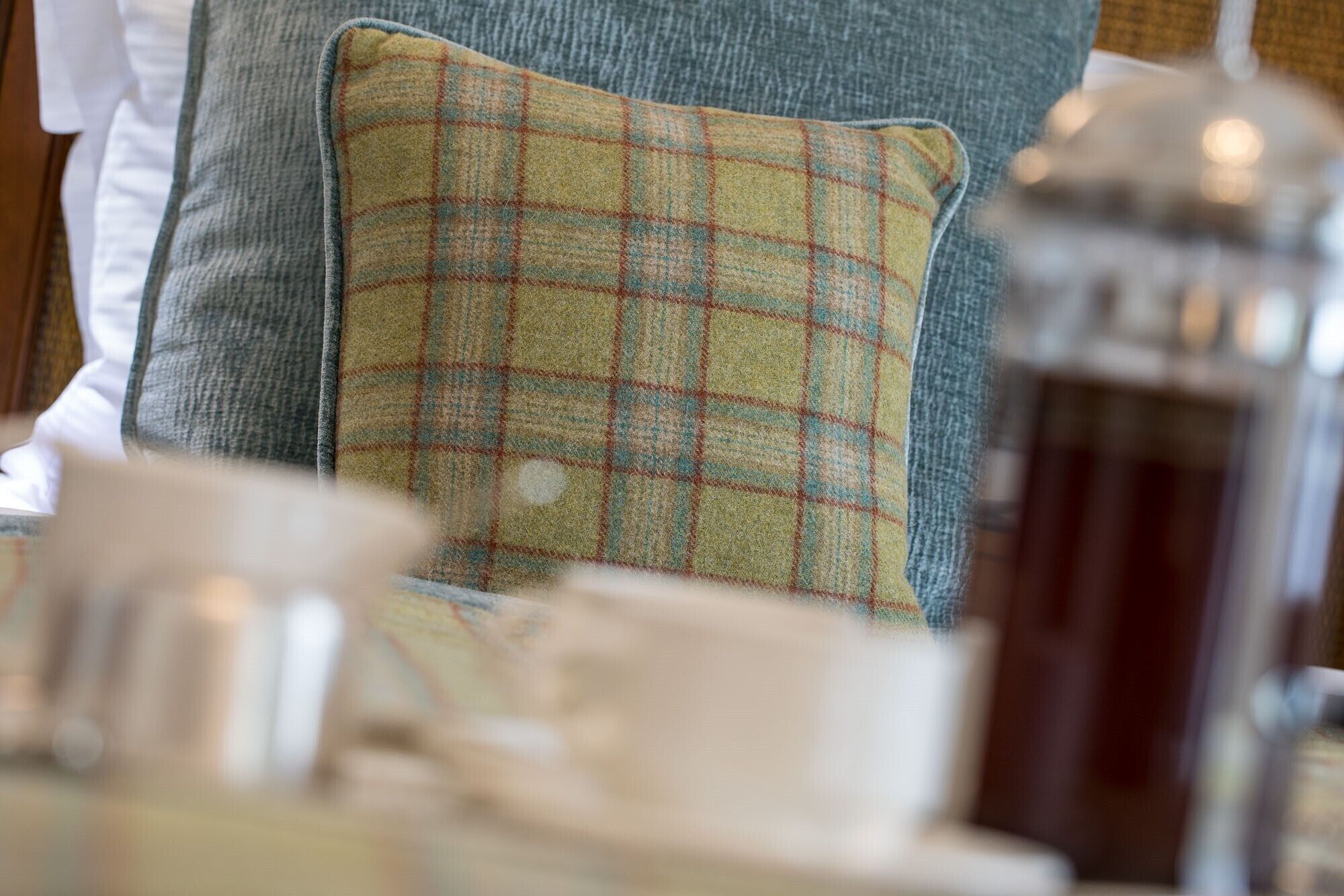 A close-up of a plaid pillow on a chair, a white cup, and a glass jar on a table.