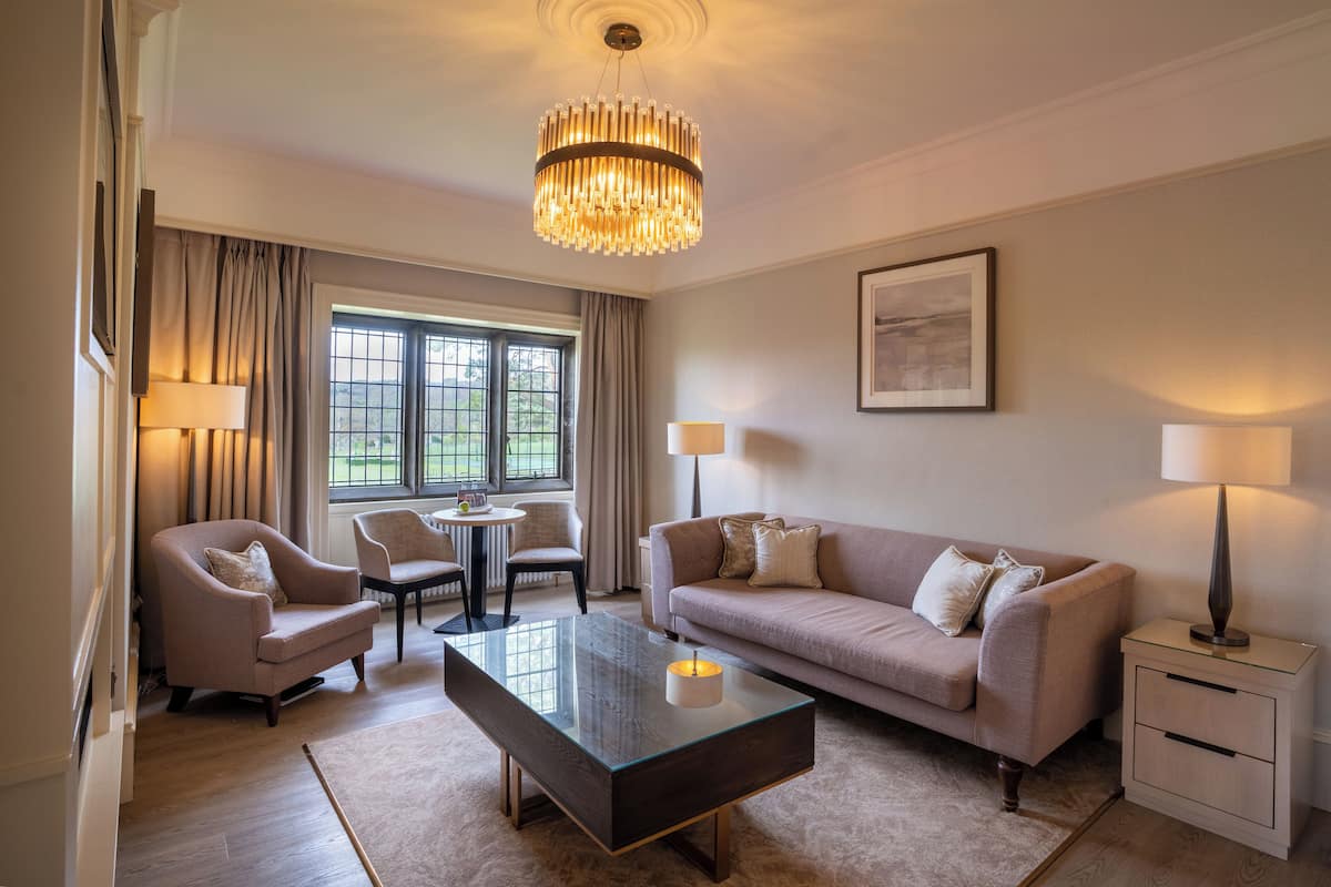 A modern hotel room with a large window, a chandelier, a sofa, a coffee table, and two lamps.