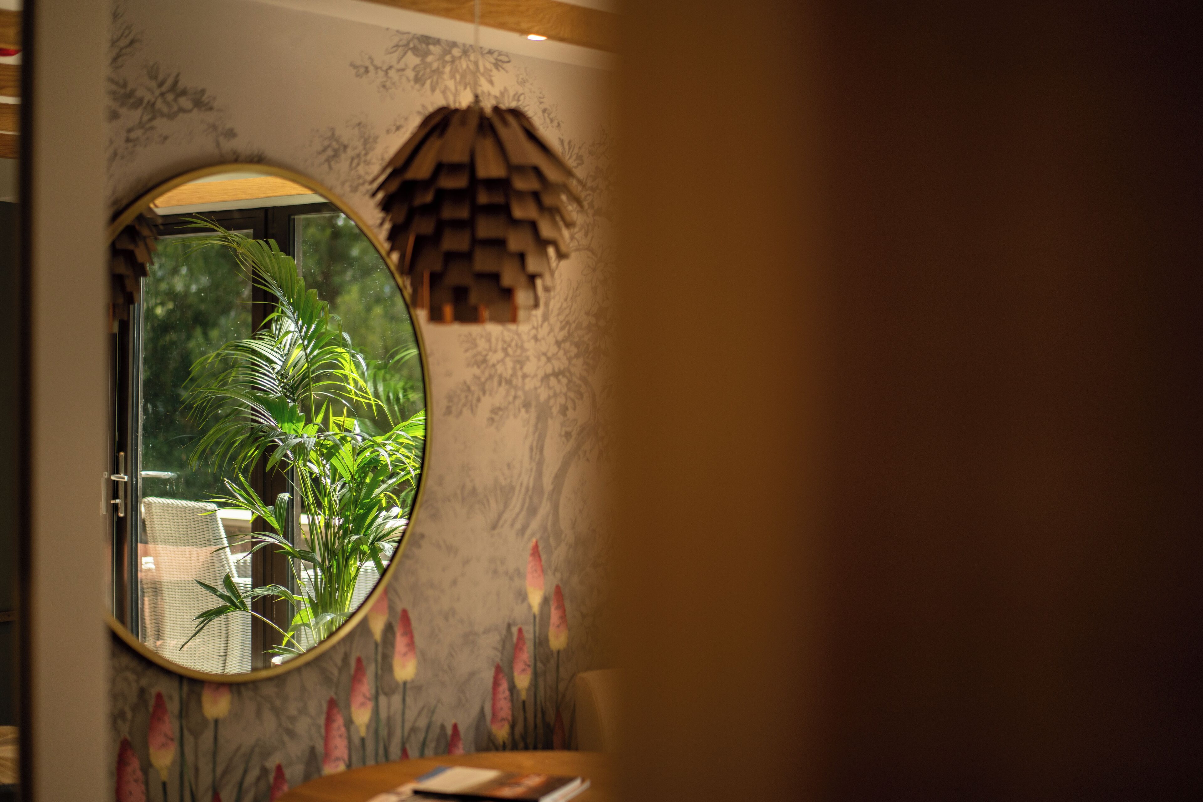 Habitació amb un mirall rodó que reflecteix una planta i una cadira, una paret decorativa amb motius florals, i una estructura de fusta que sembla una cabana.