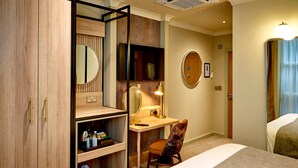A hotel room with a wooden wardrobe, a desk with a lamp, a chair, a bed, and a round mirror on the wall. - Belmore (Sale)