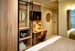 A hotel room with a wooden wardrobe, a desk with a lamp, a chair, a bed, and a round mirror on the wall.