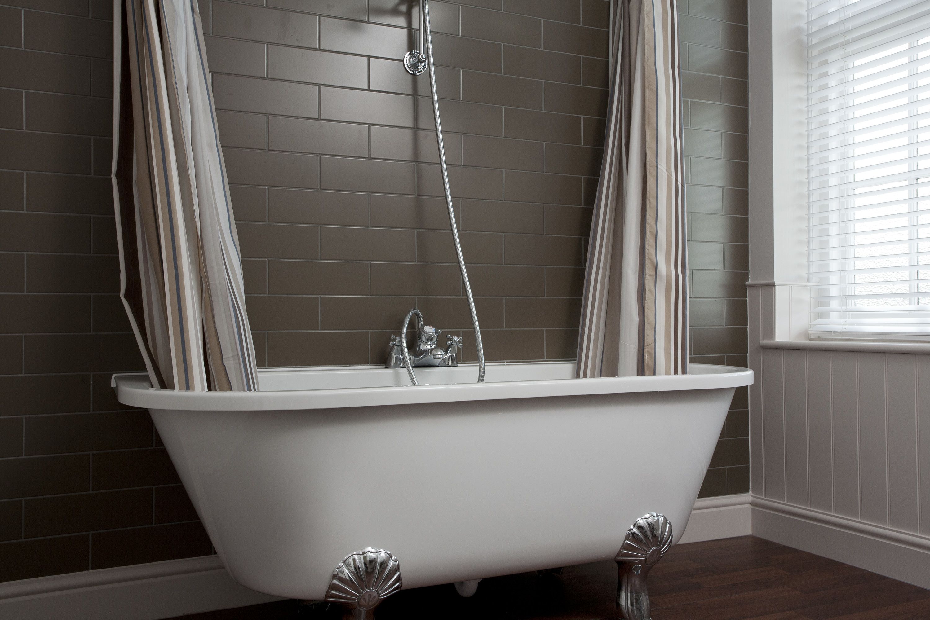 Una bañera independiente en un baño con pared de azulejos y una ventana con persianas.