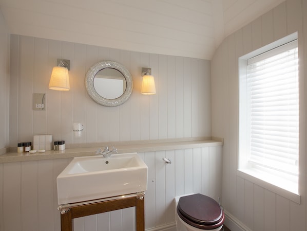 Un baño con lavamanos, tocador y espejo blancos. Hay dos luces empotradas en la pared, una ventana con persianas y un taburete de madera.