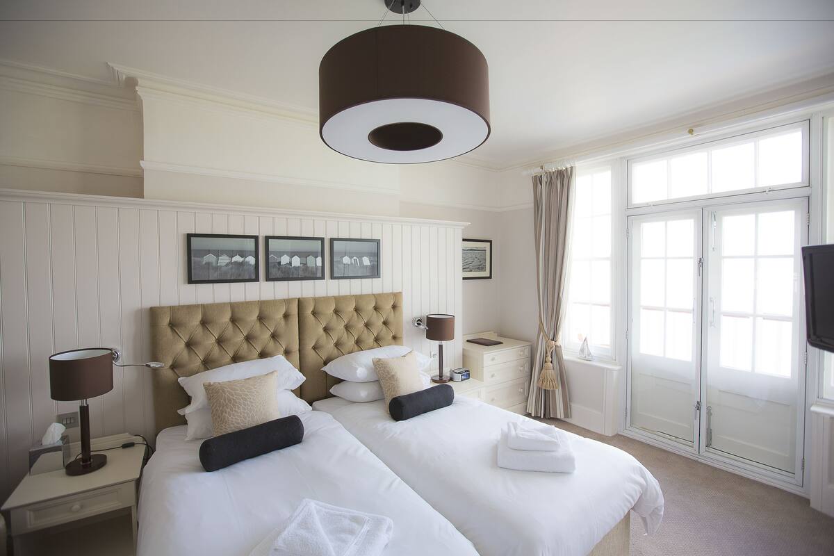 A hotel room with a large bed, two bedside tables, a modern chandelier, and large windows with curtains.