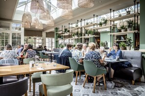 A spacious indoor dining area with high ceilings, wicker pendant lighting, and numerous potted plants on shelves. - Marine Hotel (Whitstable)