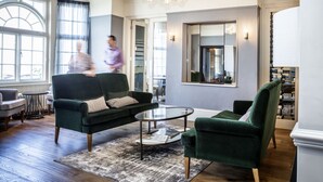 A spacious living room with a green sofa, a glass coffee table, and a chandelier. There is a large mirror on the wall and a bookshelf filled with books. - Marine Hotel (Whitstable)