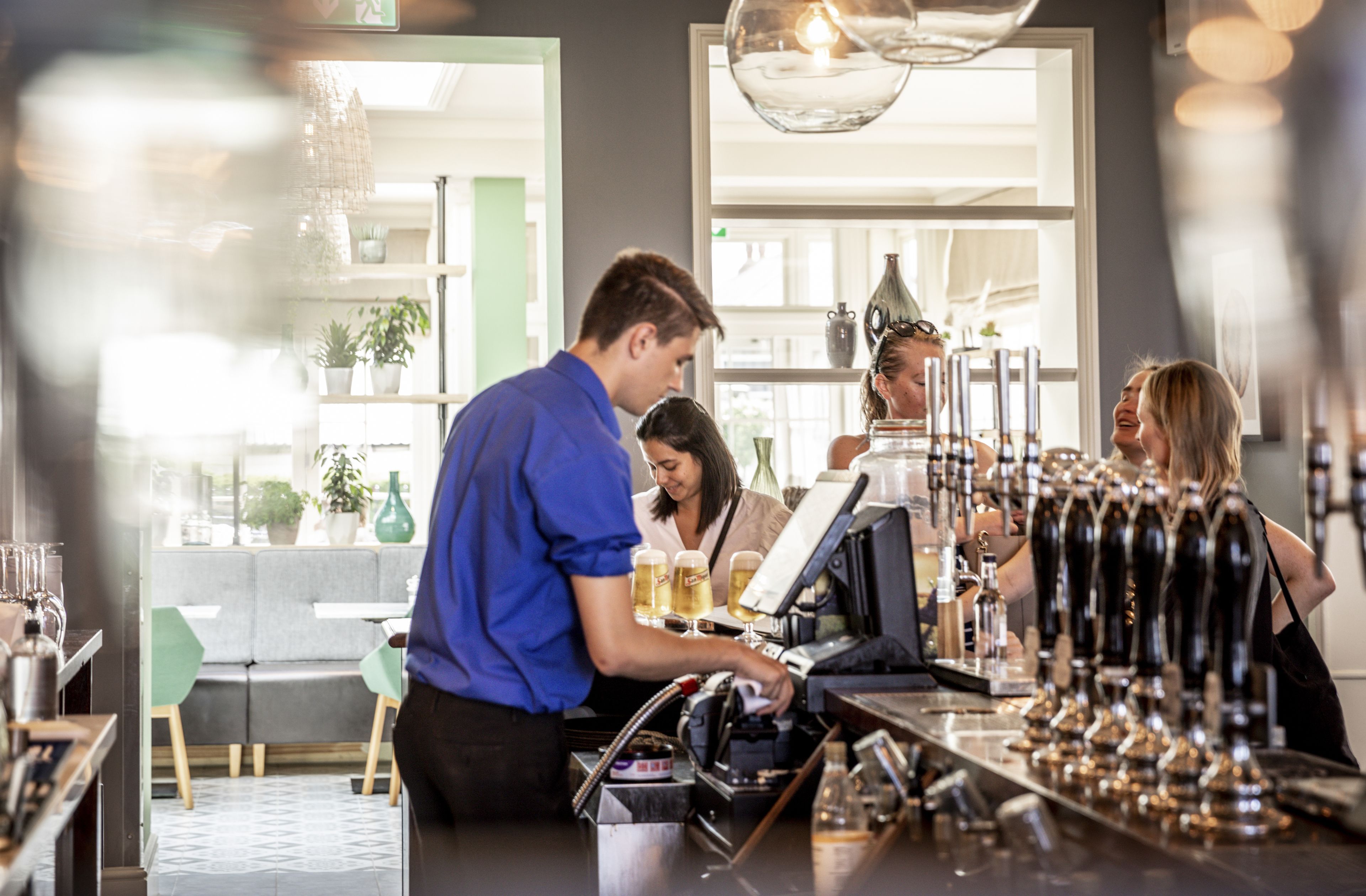 A bar area with a bartender preparing drinks, a counter with taps, and a seating area with chairs and a sofa.