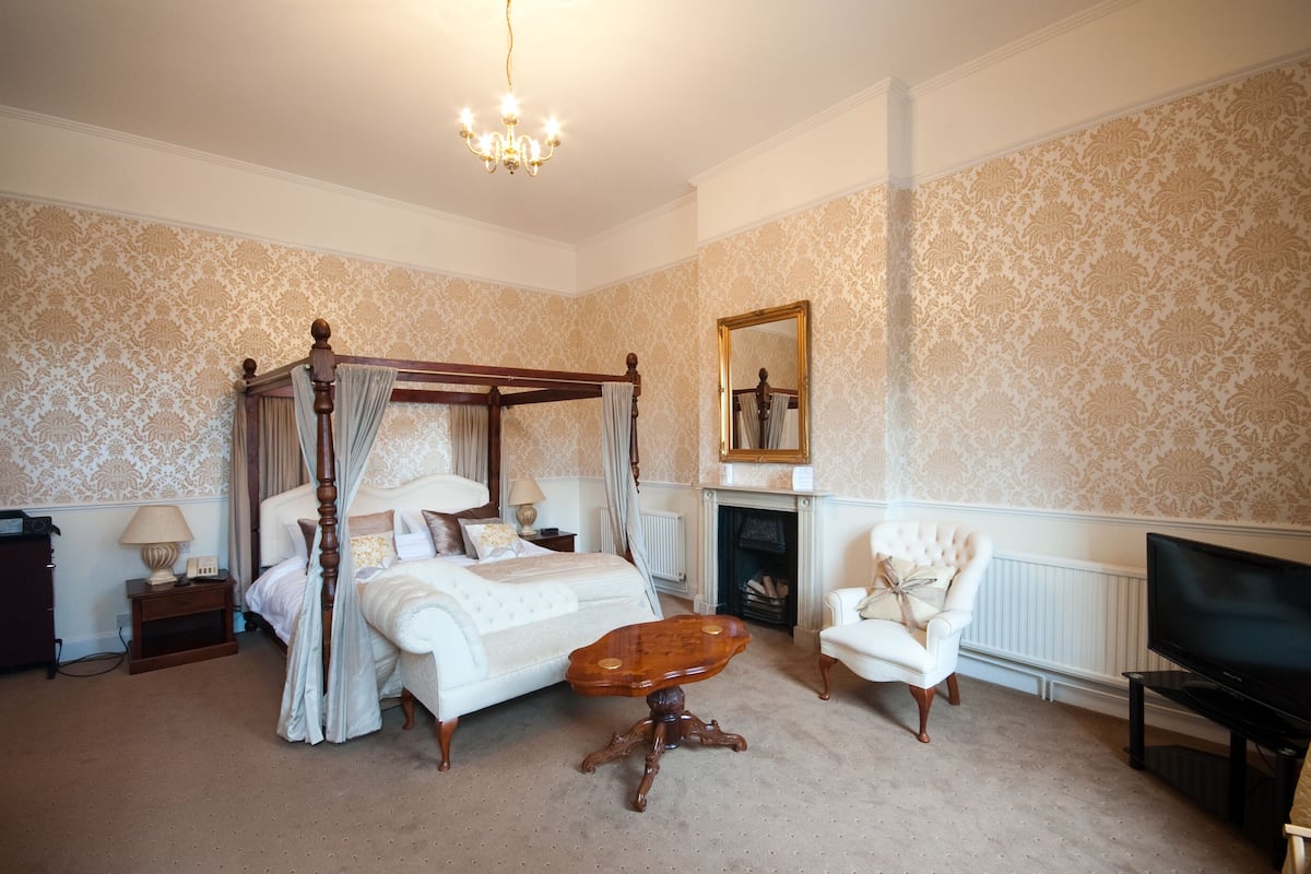 A four-poster bed with a canopy, a wooden coffee table, a white armchair, a television, and a fireplace.