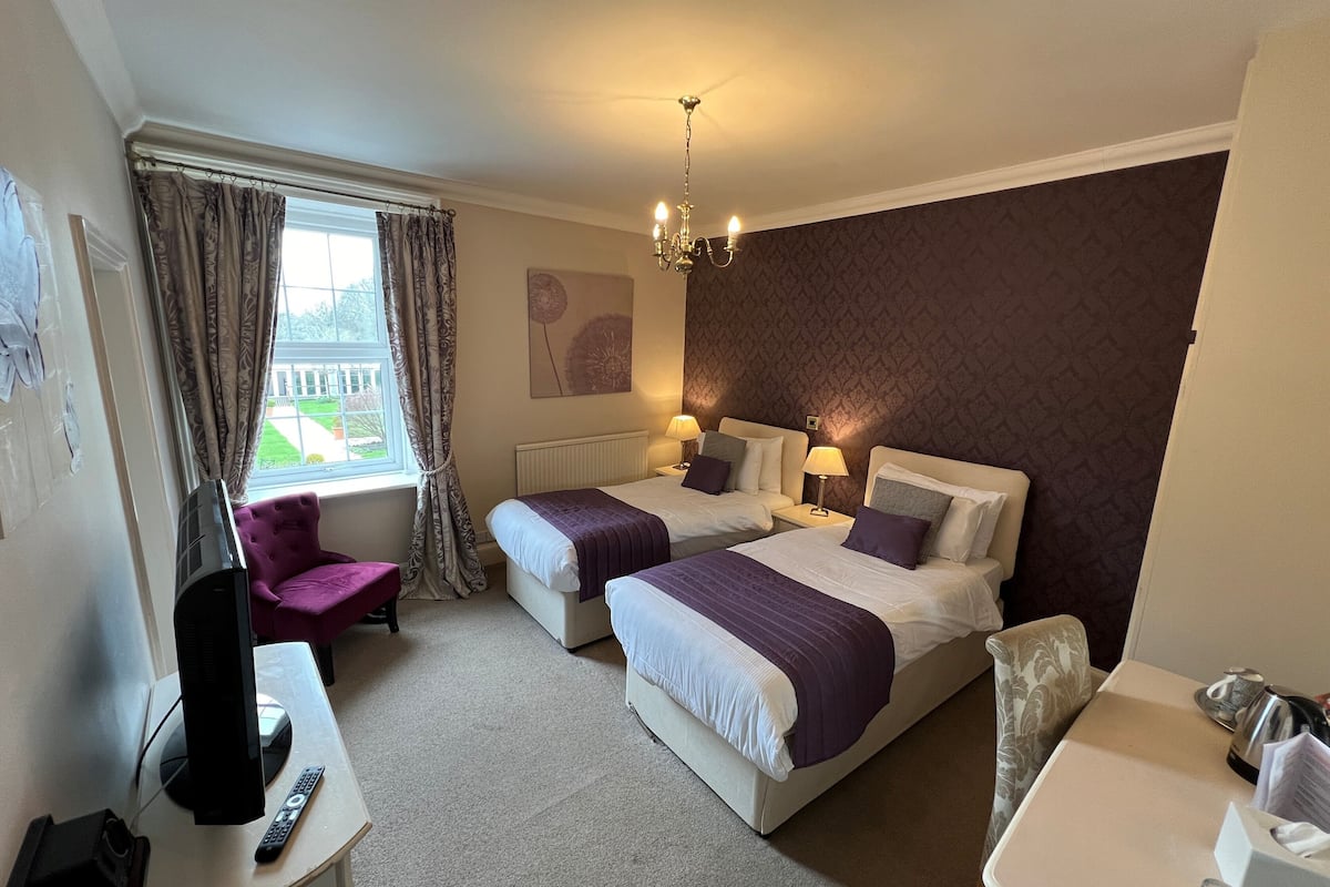 A hotel room with two beds, a desk with a computer and remote, a chair, and a window with curtains.