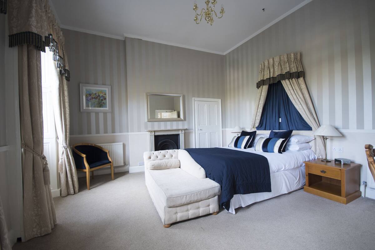 A bedroom with a large bed, a nightstand, a chair, a fireplace, and a mirror on the wall.