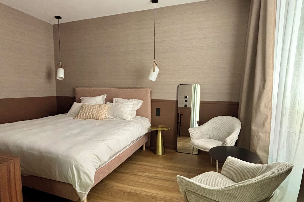 A hotel room with a large bed, a chair, a small table, and a mirror.