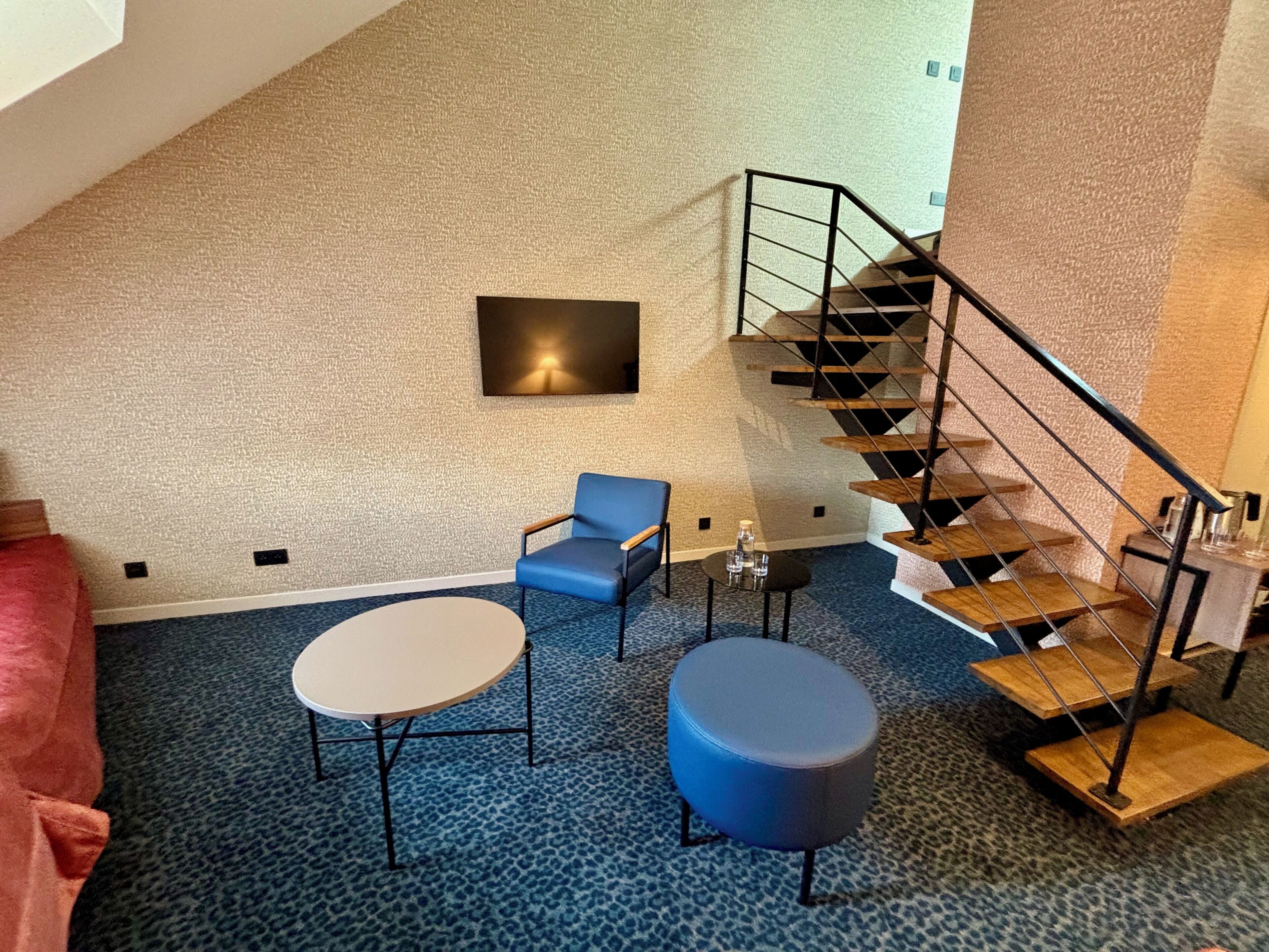Una habitación con alfombra azul, una mesa redonda, una silla azul, un reposapiés redondo, una lámpara fijada en la pared y una escalera de madera.