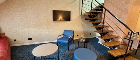 A room with a blue carpet, a round table, a blue chair, a round ottoman, a wall-mounted light, and a wooden staircase.