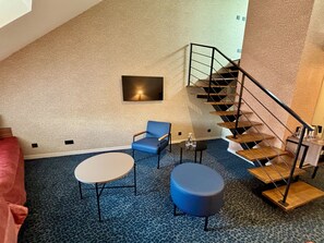 Una habitación con alfombra azul, una mesa redonda, una silla azul, un reposapiés redondo, una lámpara fijada en la pared y una escalera de madera.