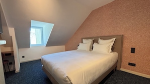 Un dormitorio con techo inclinado, una cama con ropa de cama blanca, una ventana pequeña y suelo cubierto de alfombra.