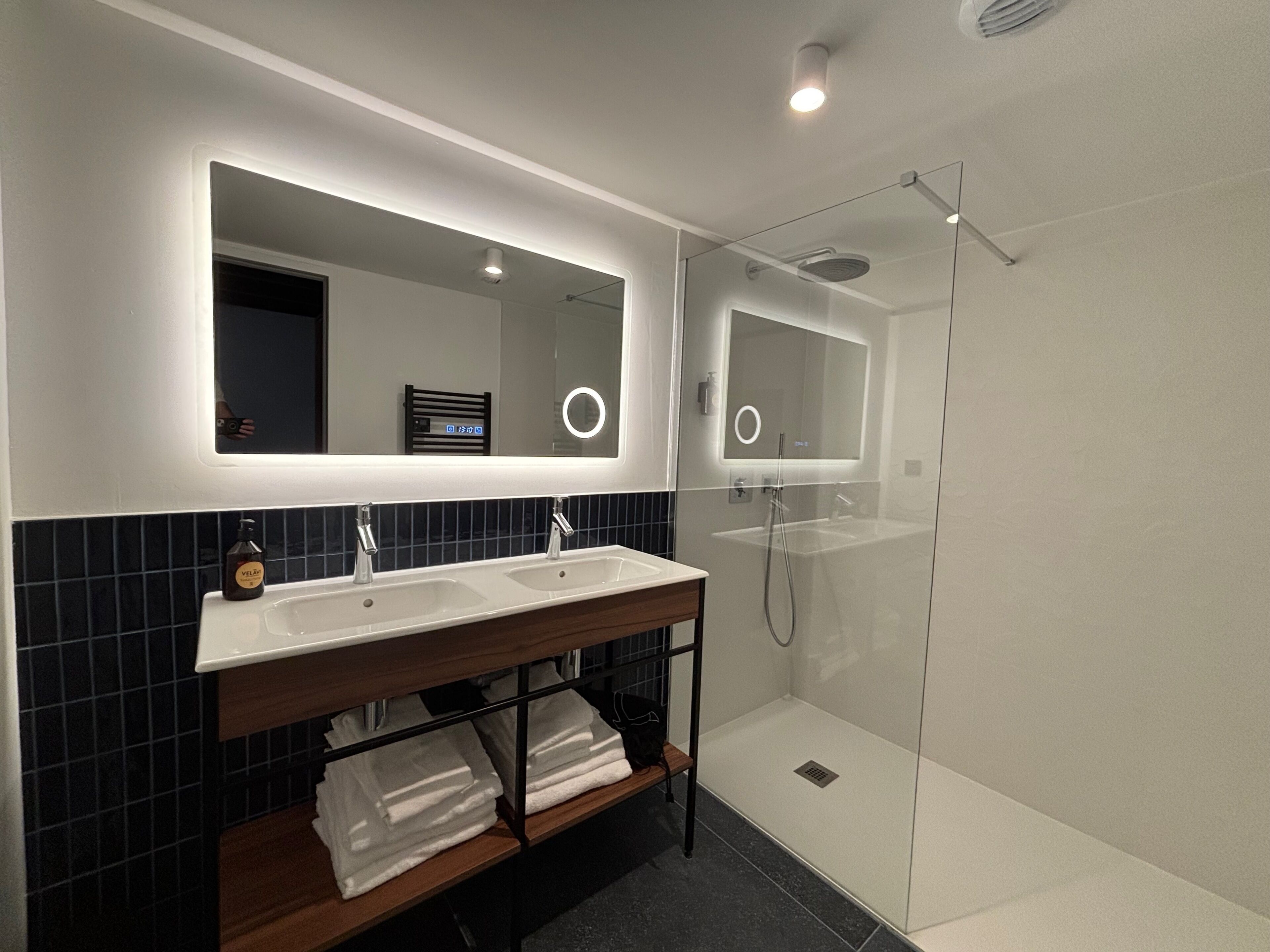 Un baño moderno con tocador doble, cabina de ducha con mampara de vidrio e iluminación integrada en el espejo.
