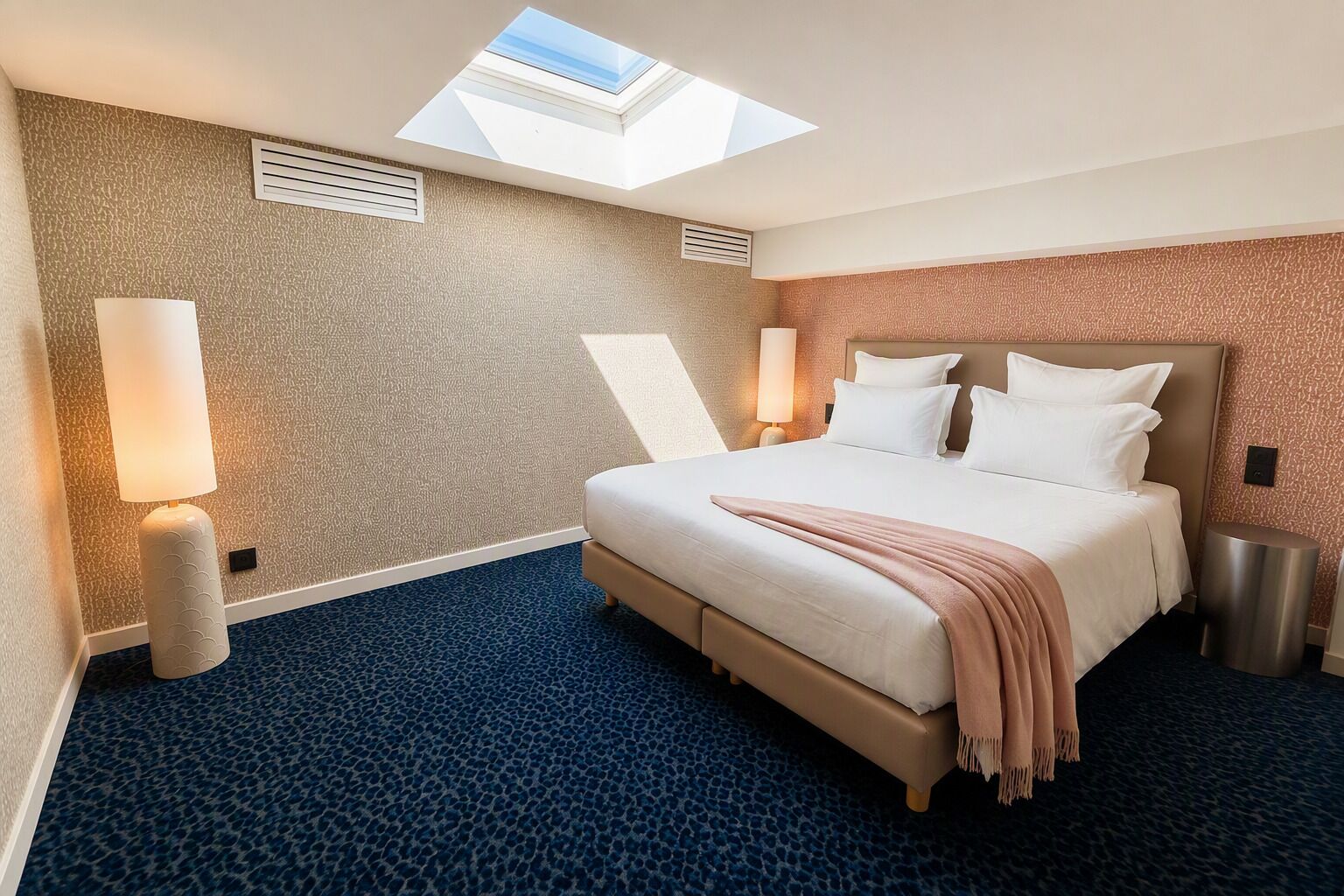 Une chambre d’hôtel avec une lucarne, un lit recouvert de draps blancs, une moquette bleue et une lampe de chevet.