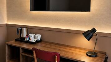 A hotel room with a wooden desk, a red chair, a black trash bin, a black desk lamp, and a wall-mounted mirror.